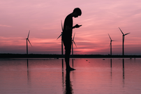 Silhouette photo of man standing while using smart phone in front og wind turbins and a pink sunset. Credits: Unsplash.com/@themodernaffliction