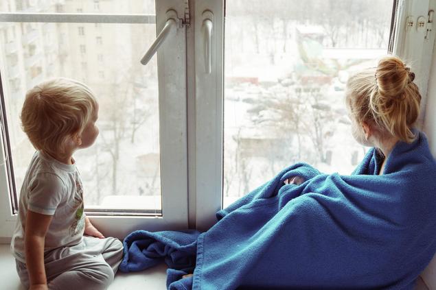 Barn med teppe rundt seg sitter i vinduskarmen og titter ut på vinterlandskap. Foto: Vitolda Klein på Unsplash.